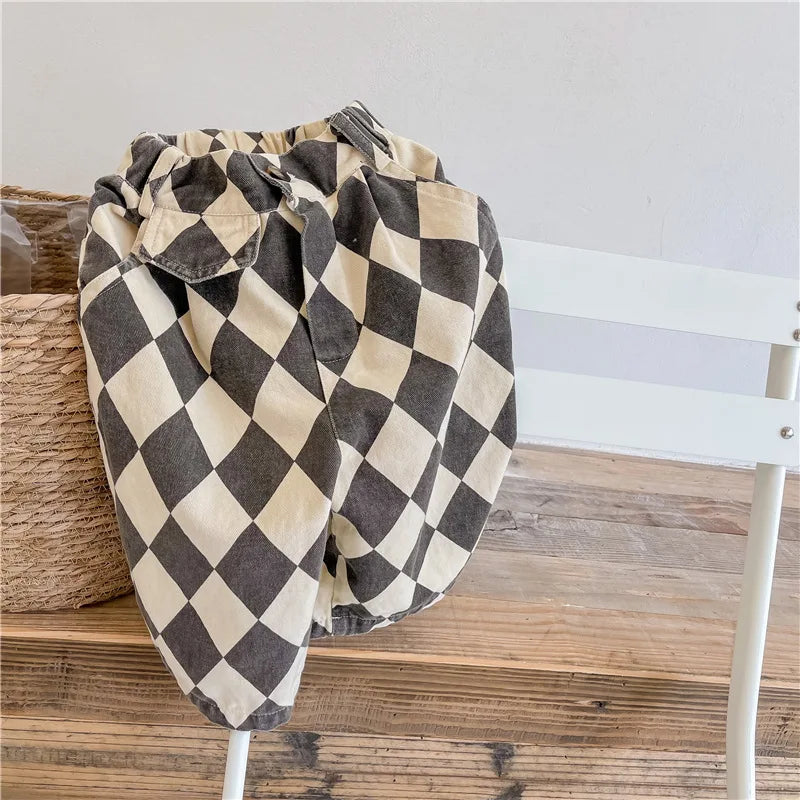 A pair of kids' washed cotton harem pants with a black and beige checkered pattern folded on a white wooden chair.