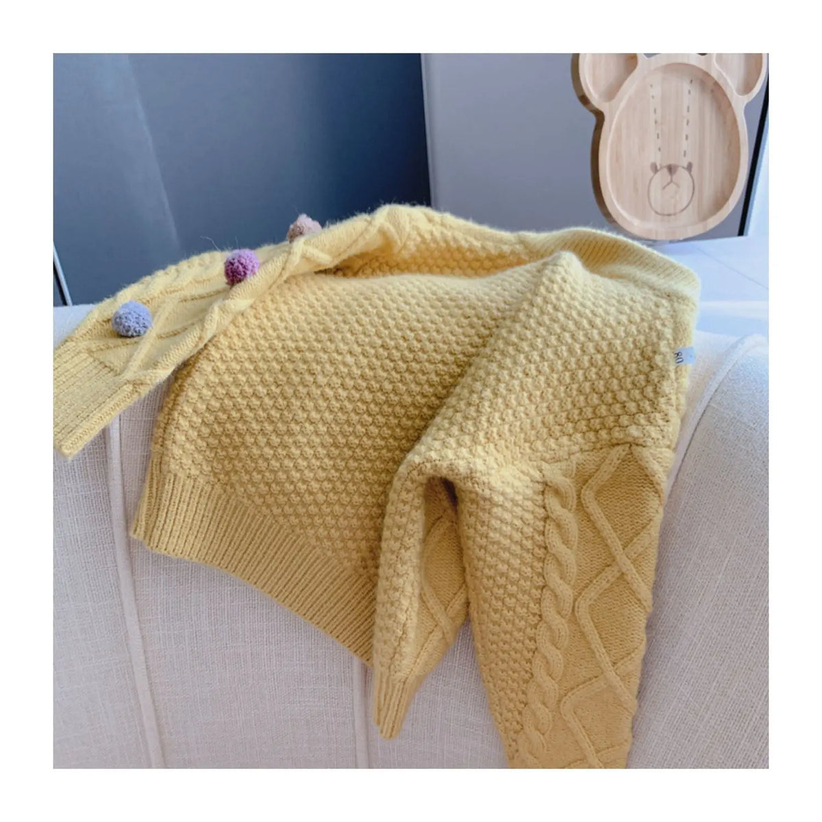 A yellow knitted cardigan sweater with a pom-pom trim is displayed on a white surface.