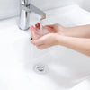 Hands are washing under a modern faucet in a white sink, with water flowing and a drain stopper visible.