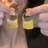 A pair of gold geometric tassel drop earrings being held up against a blurred background.