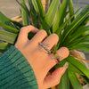 Hand mit Regenbogen-Perlen-Drehring an einem Finger, vor grünem Blätterhintergrund.