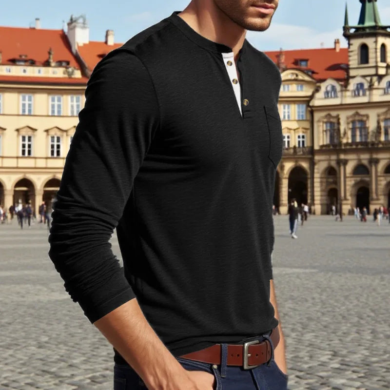 A man wearing a black long-sleeve shirt with a chest pocket and dark jeans standing in a historic European square.
