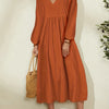 A woman wearing a rust orange cotton-linen blend dress with a V-neck and bell sleeves, standing against a light wall.