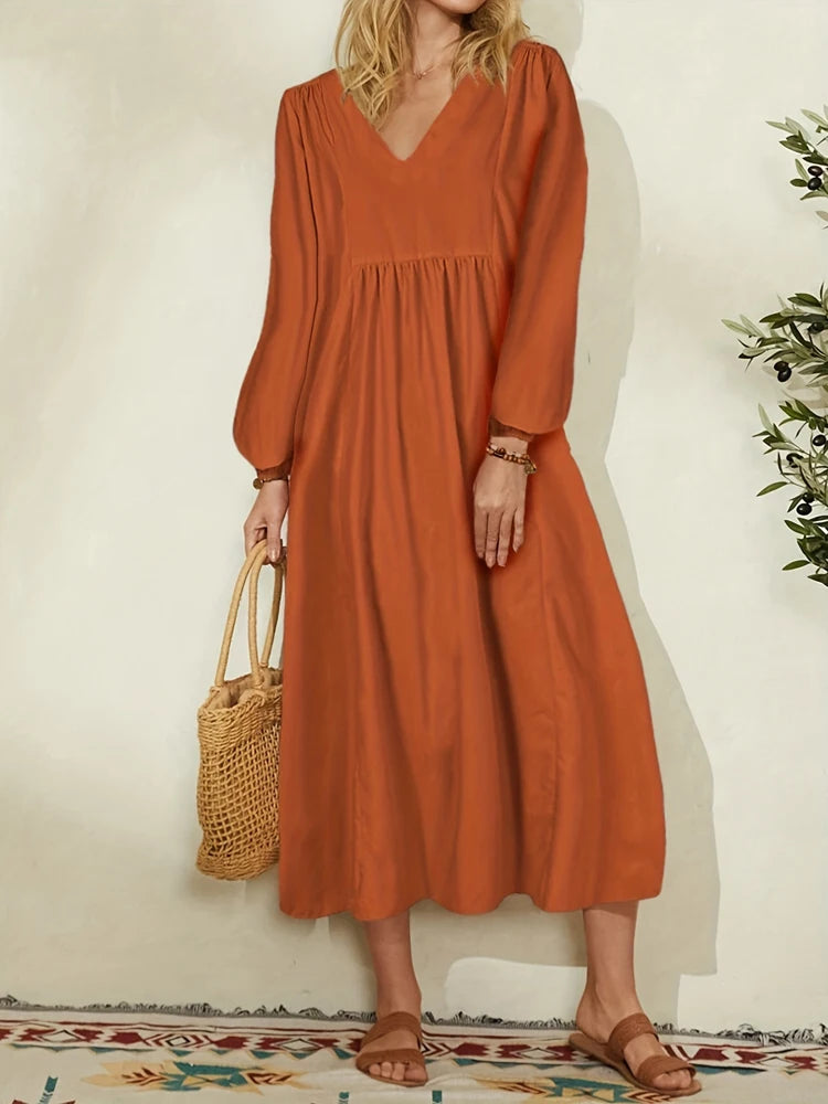 A woman wearing a rust orange cotton-linen blend dress with a V-neck and bell sleeves, standing against a light wall.