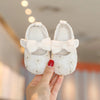 A hand holds a pair of white baby first walker shoes with pink bows.