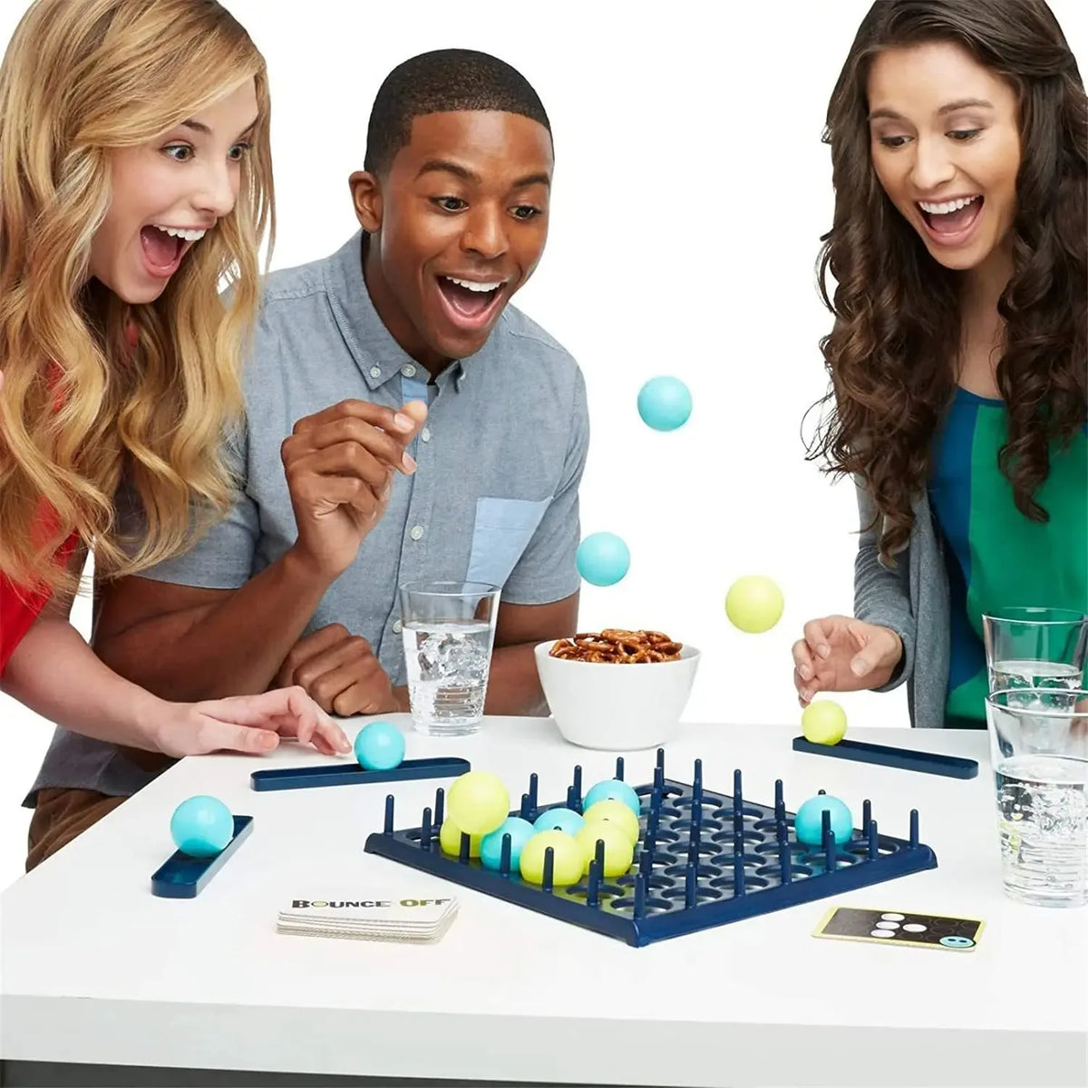 Three people laughing while playing a blue and yellow bounce ball tabletop game on a white table.
