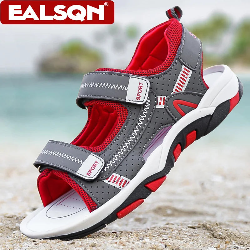A pair of gray and red boys' breathable summer sport sandals with adjustable straps resting on a sandy beach.