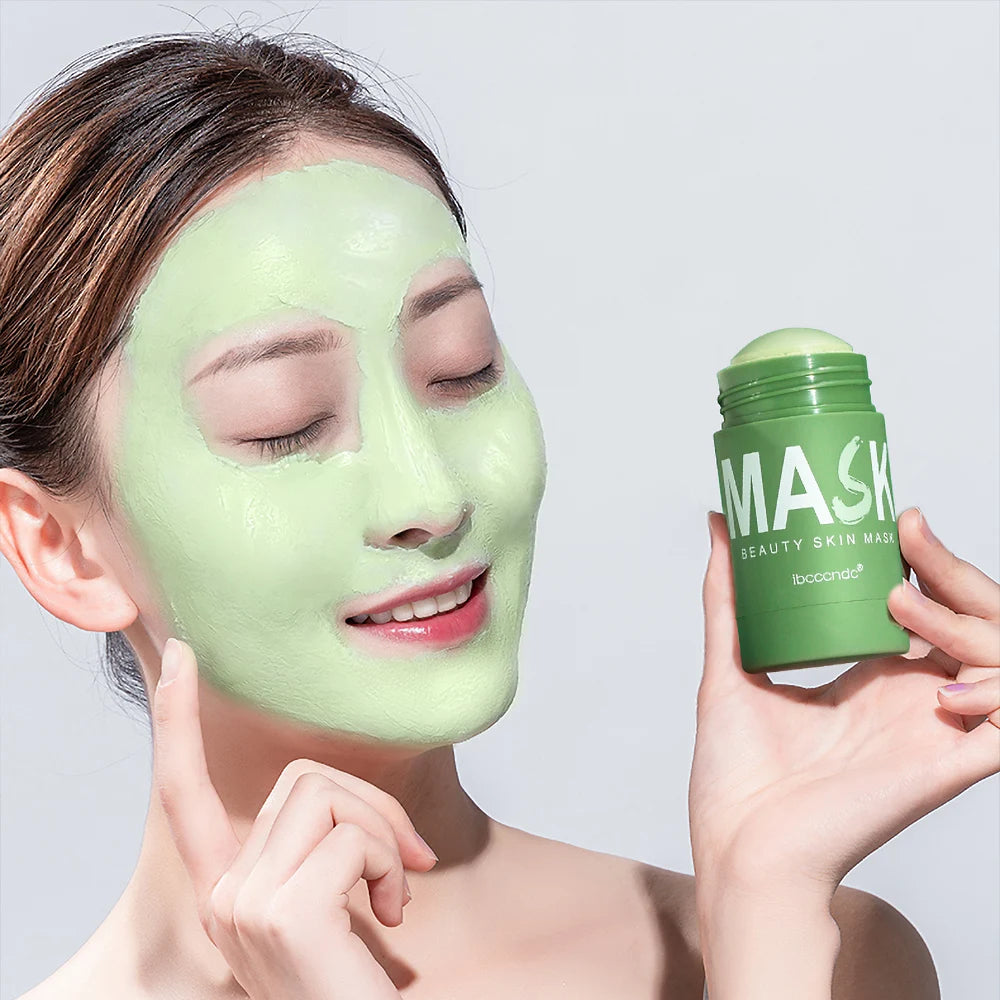 A woman applies a green facial mask to her face while holding a jar of mask product.