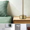 A gold-toned desk lamp with a white dome shade sits on a wooden surface next to a green cushion.