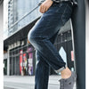 A man wearing dark denim jeans and a plaid shirt stands outdoors, modeling leather slip-on loafers.