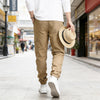 A man wearing tan cargo pants and a white shirt walks down a city street while holding a straw hat.
