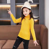 A young girl wearing a yellow knit turtleneck sweater and dark jeans, posing with one finger raised.