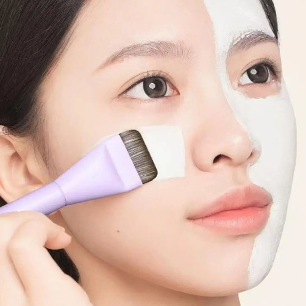 A woman applies a white facial mask using a professional flat foundation brush on her face.