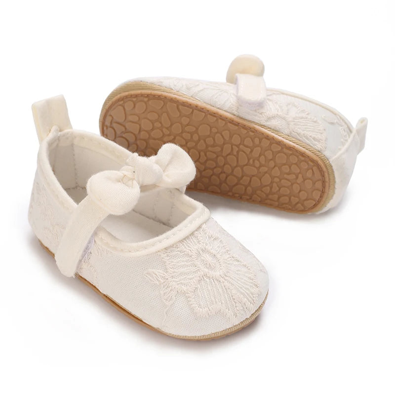 A pair of soft non-slip baby first walker shoes in off-white fabric with a tan sole and a decorative bow detail.