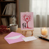A pink card featuring a red heart tree silhouette with a couple standing beneath it, placed on a wooden table.
