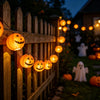 A wooden fence decorated with string lights featuring glowing orange pumpkin lanterns in a dark garden setting.