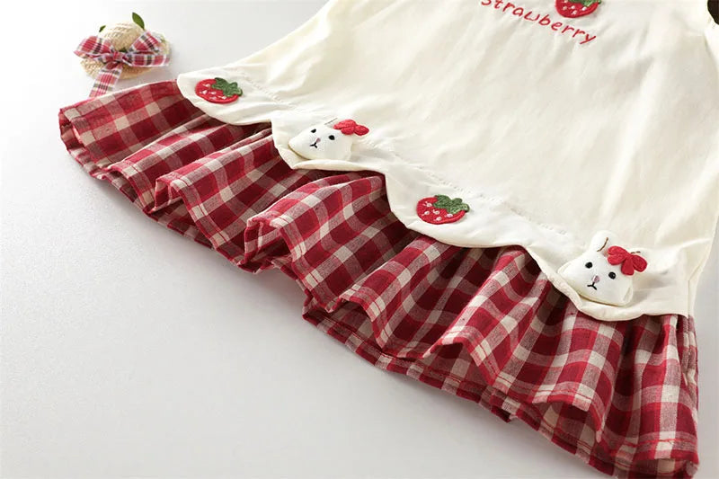A cream summer pinafore dress featuring a red plaid ruffle and strawberry and bunny print details.