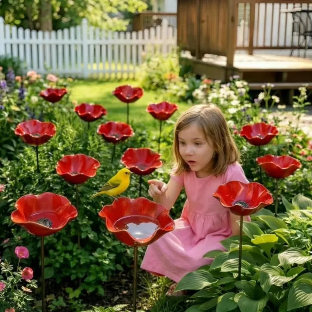 A young girl sits among red metal flowers in a garden with a white picket fence and wooden structure.