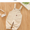 A beige cotton toddler overalls laid flat on a wooden surface next to a small camera.