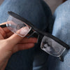 A hand holds a pair of black frame reading glasses with clear lenses against a blue denim background.
