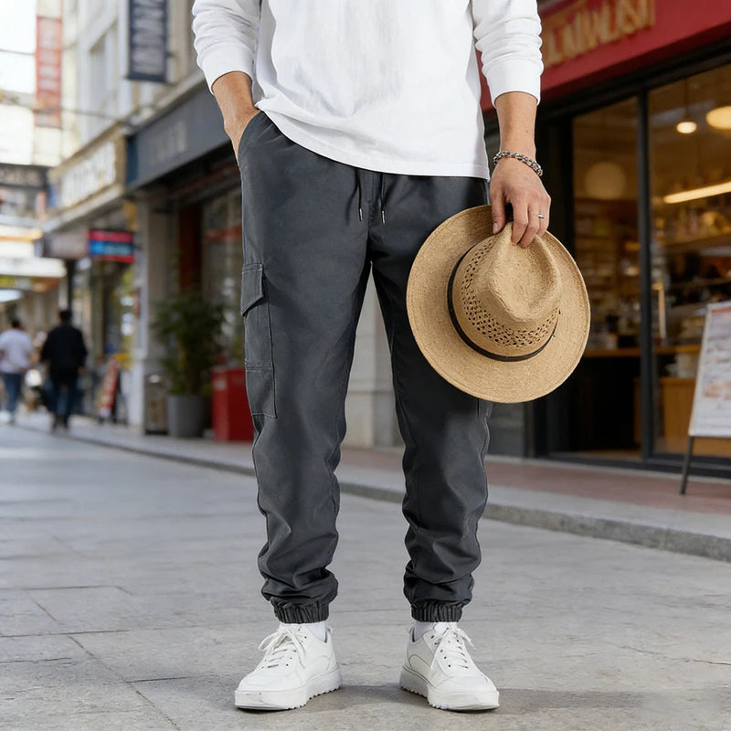 A man wearing dark gray casual cargo pants and a white shirt stands on a sidewalk.