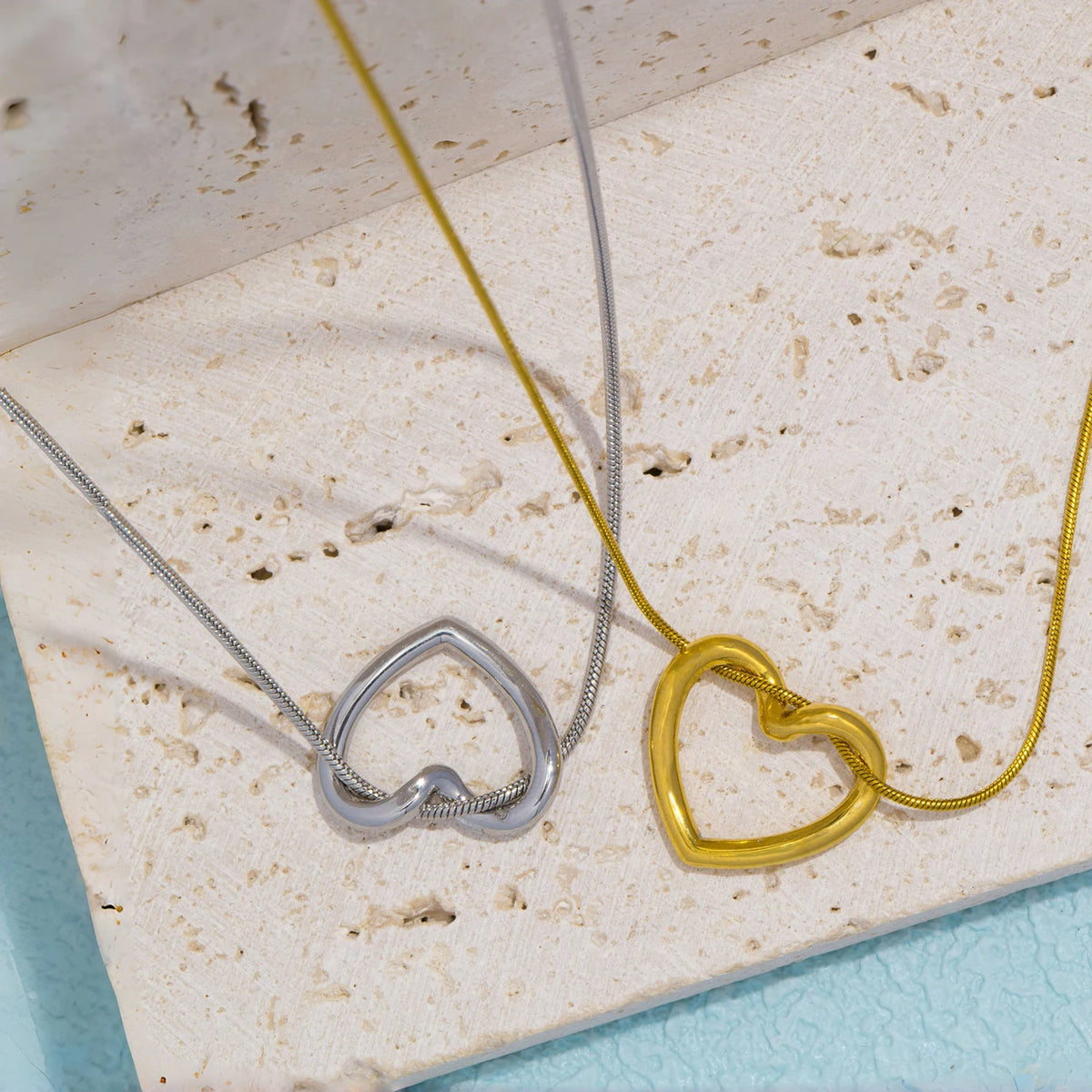 A dainty gold heart pendant necklace with a snake chain resting on a light beige surface.