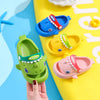 A hand holds a green kids shark clog sandal with a red accent and anti-slip sole on a yellow background.