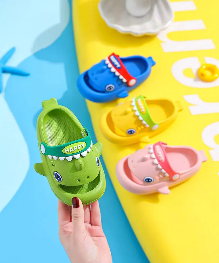 A hand holds a green kids shark clog sandal with a red accent and anti-slip sole on a yellow background.
