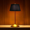 A modern table lamp with a black fabric shade and a gold base sits on a wooden surface against a warm orange background.