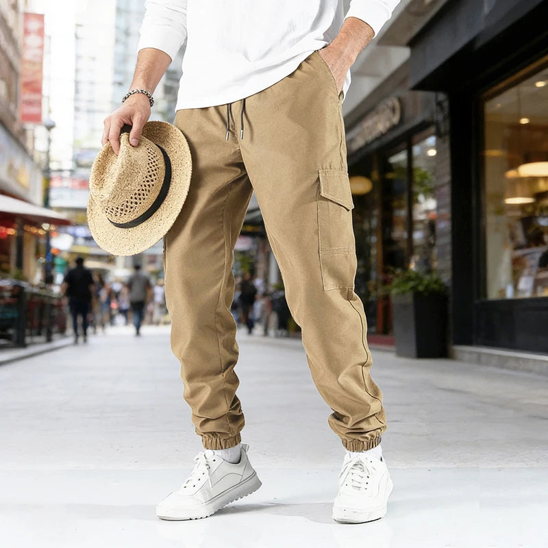 A man wearing tan cargo pants and a white shirt walks outdoors while holding a straw hat.