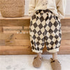 A child wearing brown and beige checkered harem pants with pockets, standing in front of a wooden step.
