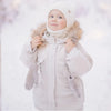 A baby wearing a beige winter down jacket and pants set with a faux fur hood in the snow.