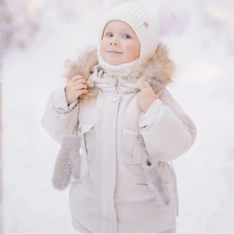 A baby wearing a beige winter down jacket and pants set with a faux fur hood in the snow.