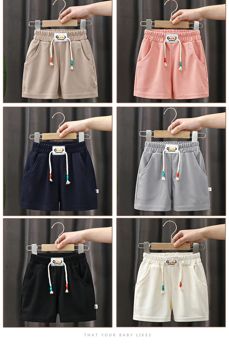 A collection of kids beach shorts in various colors including beige pink navy gray black and cream shown on hangers.