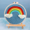 Kit de bordado con aguja mágica con diseño de arcoíris colorido sobre fondo azul, montado en soporte de madera.