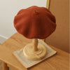 A terracotta orange wool beret hat displayed on a wooden stand.