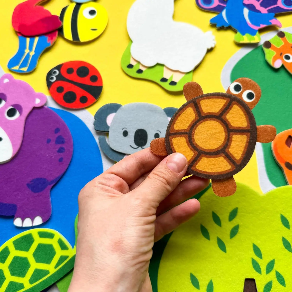 A hand is holding a felt zoo animal, specifically a turtle, against a colorful felt story board background.