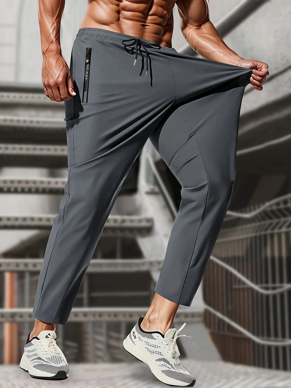 A muscular man wearing gray high-stretch joggers with black laces and white athletic shoes.