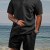 A man wearing a black jacquard short-sleeve shirt and drawstring shorts set standing near the water.