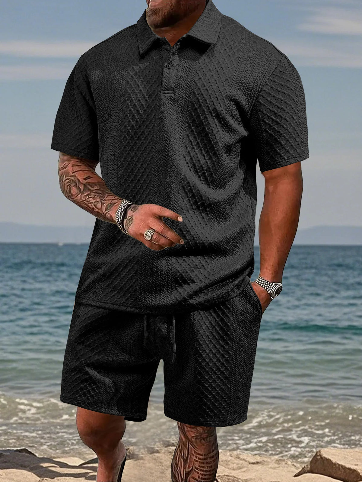 A man wearing a black jacquard short-sleeve shirt and drawstring shorts set standing near the water.