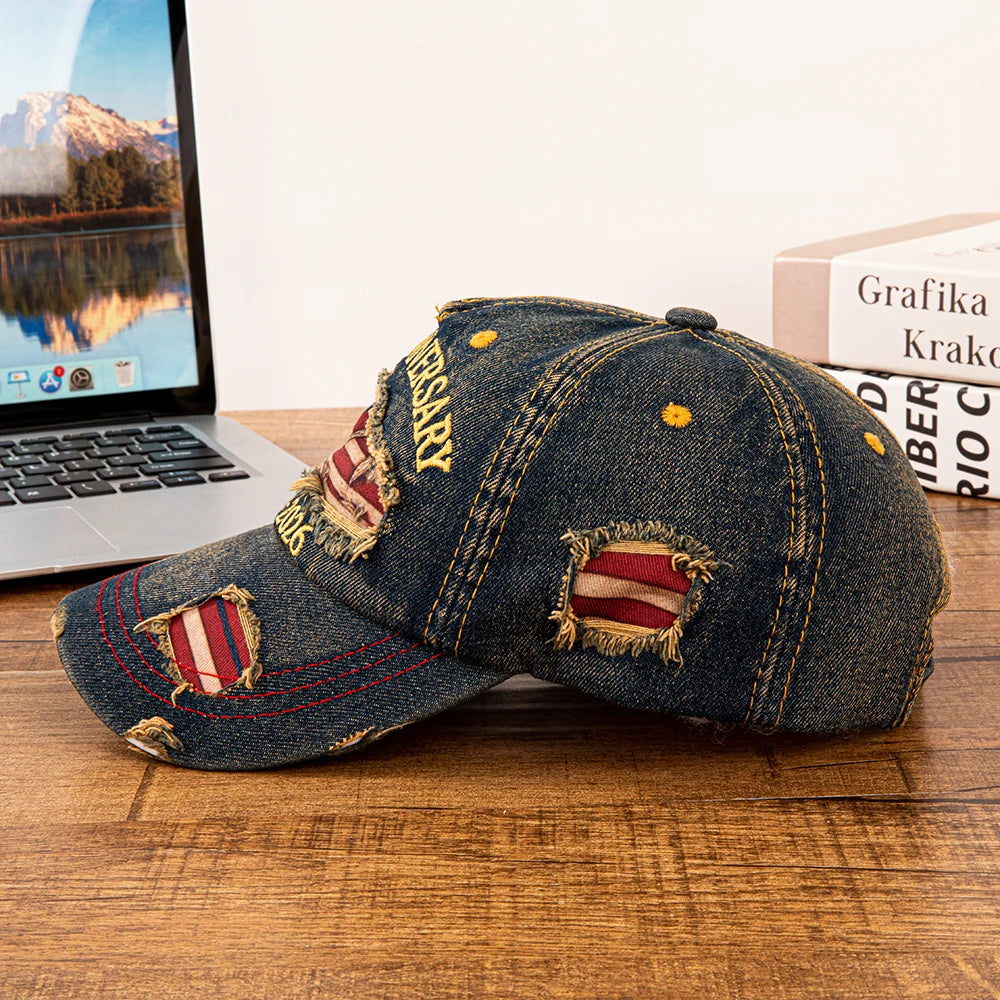 Mørk denim-baseballcap med slidt flagdesign og tekst, hvilende på træoverflade ved siden af en bærbar computer.