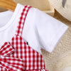 A white t-shirt with a red plaid apron worn by a child, resting on a beige knitted surface.