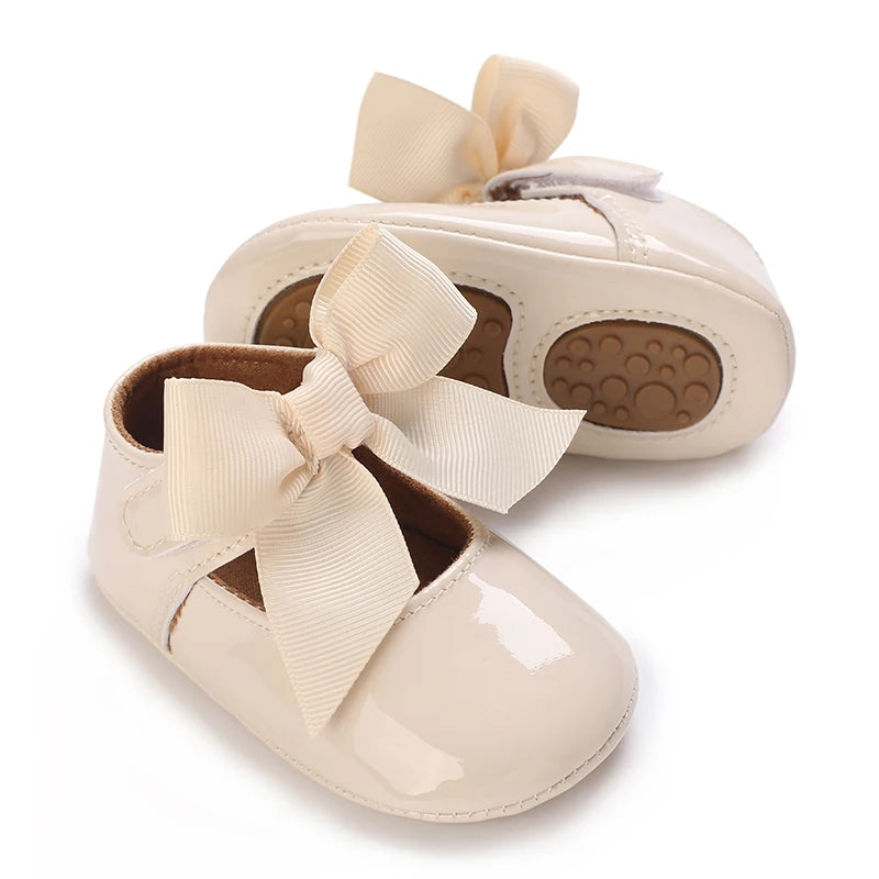A pair of beige soft non-slip baby first walker shoes with satin bows.