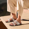 A pair of beige women's cork footbed slide sandals with white straps resting on a light wooden surface.