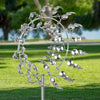 Escultura cinética de jardín de un molino de viento metálico plateado con aspas delicadas y reflectantes sobre césped verde.