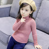 A young girl wearing a pink knit turtleneck sweater and a beige knit hat sitting on a gray sofa.