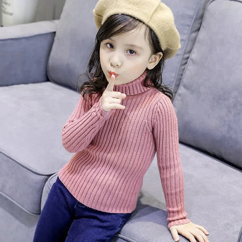 A young girl wearing a pink knit turtleneck sweater and a beige knit hat sitting on a gray sofa.