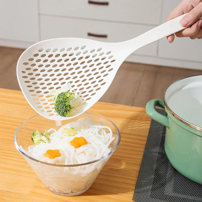A hand is holding a white long-handled kitchen strainer spoon over a glass bowl filled with clear noodles.