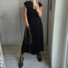 A woman wearing a black sleeveless pleated midi dress and a black handbag standing in a well-lit hallway.