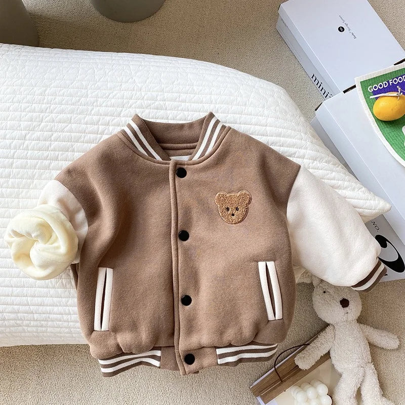 A brown corduroy jacket with white trim and a teddy bear patch is displayed on a white textured surface.